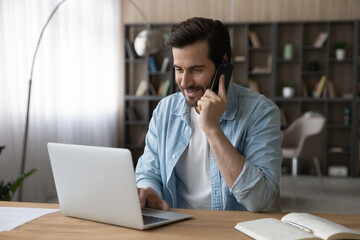 Smiling businessman talking on smartphone, using laptop, sitting at desk, friendly manager...
