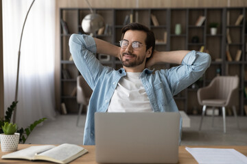 Positive dreamy businessman in glasses sitting at desk with laptop, leaning back, stretching hands, relaxing after work finished and daydreaming about good future, new career opportunities