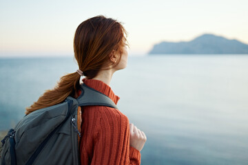 woman tourist in an orange sweater and with a backpack on her back is resting near the sea and mountains in the distance