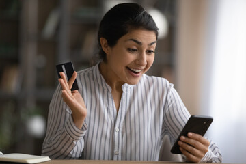 Close up excited Indian woman holding credit card, using smartphone, reading good news, checking balance, surprised customer received money refund or great shopping offer, making internet payment