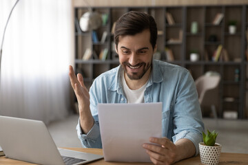 Excited businessman reading good news in letter, notification, sitting at workplace with laptop, overjoyed young man holding documents, received money refund or job promotion, celebrating success