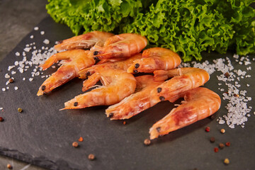 Fresh shrimps lie with lettuce leaves and lemon on a black slate board, on a dark gray table, sea salt is poured around the shrimps. The concept of healthy food, fresh sea products