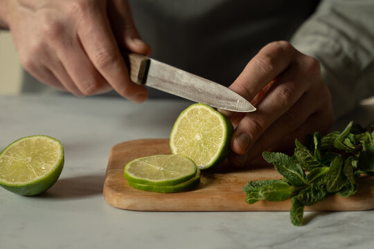 Bartender is cutting a juicy green lime on a chopping board with a knife, and a sprig of mint is lying next to it.