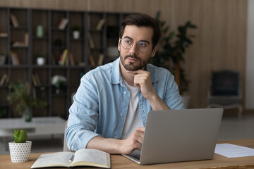 Thoughtful dreamy businessman in glasses sitting at work desk with laptop, looking to aside,...