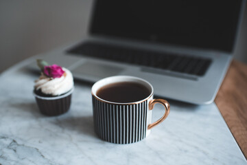 Cup of fresh black tea with creamy cupcake, open laptop on  mable coffee table close up. Good morning.