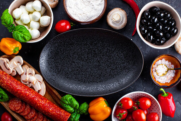  Pizza ingredients on the dark background and black plate. Pepperoni sausage, mozzarella cheese, tomatoes, olives, mushrooms and flour are different products for making pizza and pasta