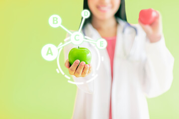 Selective focus and close up to hand holding green apple with mixed vitamin icons. Blur female nutrition doctor on isolated light green background. Healthy and Vitality Concept.