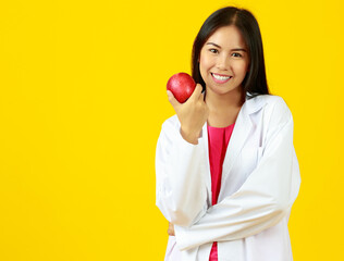 Asian beautiful female doctor wearing white gown uniform, smiling and looking at camera, crossing arms and holding red apple to present nutrition, vitamin and healthy with isolated yellow background.