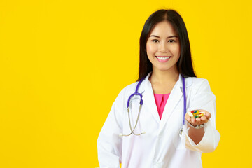 Asian young female doctor wearing white gown uniform with stethoscope, smiling, holding, presenting vitamin capsule pills with isolated yellow background and copy space. Nutrition, Healthy Concept.