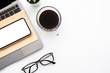 Fototapeta premium Smartphone with blank mockup screen is on top of laptop computer over white office desk table with supplies. Top view with copy space, flat lay.