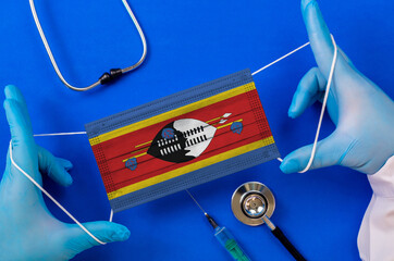 Medical mask with the flag of Swaziland in the hands of a doctor and stethoscope, flat lay....