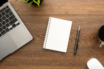 Blank notebook is on top of wood office desk table with laptop computer, and office supplies. Top view, flat lay.