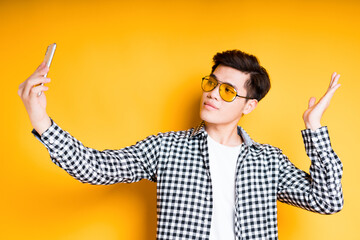 Handsome asian in sunglasses stands on a yellow background and poses sharing a selfie on the phone