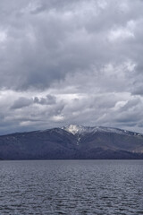 波立つ湖の向こうに見る曇り空の下の残雪の山。