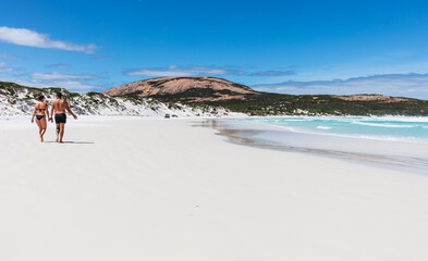 A young couple stoll along on the prisitine white sand beach and sparkling waters of Wharton Bay in the Cape LeGrande National Park on a clear summer day. - obrazy, fototapety, plakaty