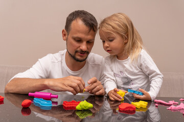 Young caucasian man plays with his daughter making colorful animals from playdough at home, happy family time together, dad and child activity, cute kid looks with interest what father does.