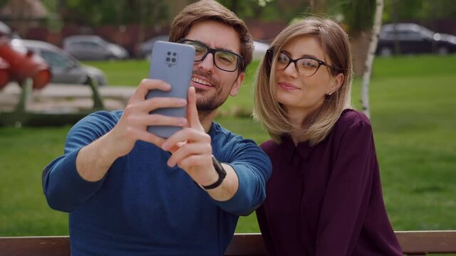 Two friends with glasses, pretty woman and handsome man sit on the bench outside take selfie with phone, feel happy smiling. Social networking outdoors. Portrait. Close up. Slow motion. 