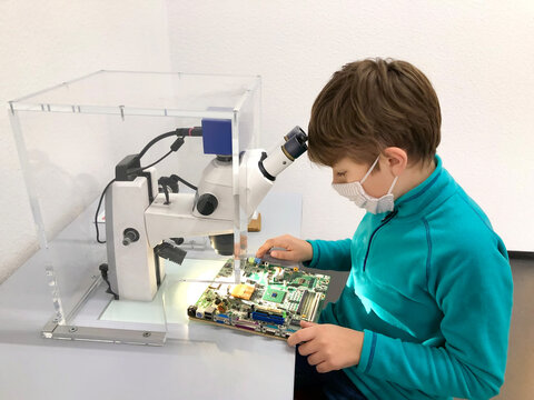 Kid boy conducts experiment with microscope in school lab. Curious inquisitive child learning physics and computer scheme. Schoolboy with medical mask due to coronavirus pandemic.