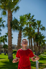 cute young teenage boy posing and having fun on the beach under palm trees in red T-shirt . Trendy boy posing. Funny and positive man in sunglasses