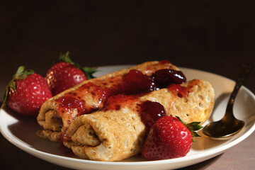 Pancakes with strawberry jam on plate. Pancakeshin rolls poured berry jam and decorated fresh strawberries on dark background. 