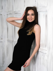 A girl is standing in a small black dress against a background of white panels, the wind blows in her face