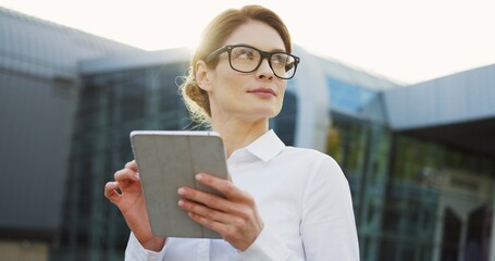 Portrait of the Caucasian good looking woman in glasses and white shirt taping on the tablet device and looking at the side near glass office building. Outdoors