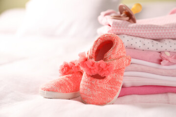 Stack of baby girl's clothes, pacifier and shoes on bed, closeup. Space for text