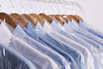 Hangers with shirts in dry cleaning plastic bags on rack against light background, closeup