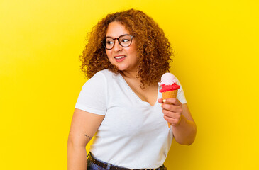 Young latin woman holding an ice cream isolated on yellow background looks aside smiling, cheerful and pleasant.