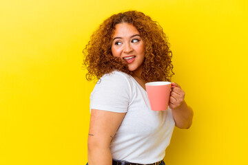 Young latin curvy woman holding a cup isolated on yellow background looks aside smiling, cheerful...