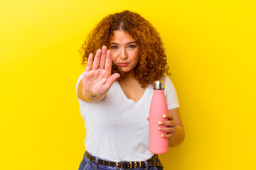 Young latin woman holding a thermos isolated on yellow background standing with outstretched hand showing stop sign, preventing you.