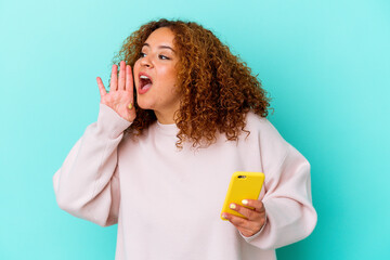Young latin woman holding mobile phone isolated on blue background shouting and holding palm near opened mouth.