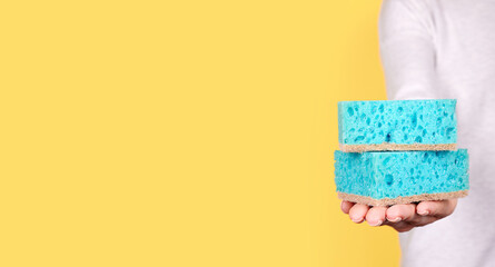 Hand with turquoise cleaning sponge on yellow background.