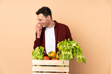 Farmer with freshly picked vegetables in a box isolated on beige background smiling a lot