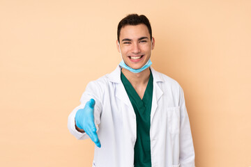Young dentist man holding tools isolated on beige background shaking hands for closing a good deal