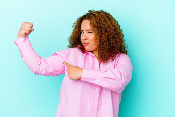 Young latin curvy woman isolated on blue background showing strength gesture with arms, symbol of...