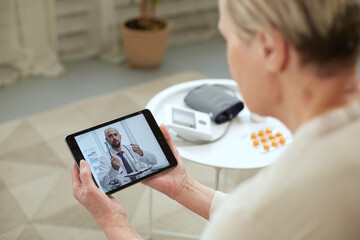 Telemedicine concept. Old woman with tablet during an online consultation with her doctor in her living room - obrazy, fototapety, plakaty
