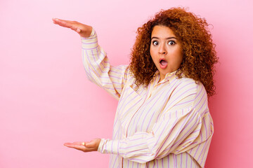 Young latin curvy woman isolated on pink background shocked and amazed holding a copy space between hands.
