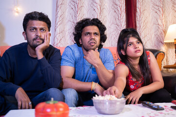 Group of young millennial friends interestingly watching live streaming sports or movie while sitting on sofa during late nigt at home.