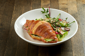 Delicious and great breakfast - croissant sandwich with salmon, cream cheese and cucumber with mixed salad, served in a white bowl on a wooden background