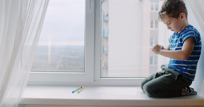 Little boy with curly hair draws on the window at home with crayons. 
