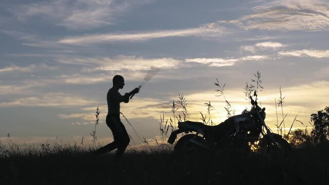Man exercising with nunchucks near a motorcycle. Silhouette at sunset, blue sky and cloudies. High quality FullHD footage