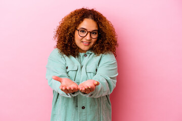 Young latin curvy woman isolated on pink background holding something with palms, offering to...