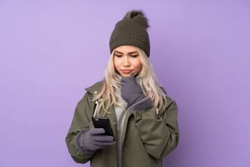 Teenager blonde girl with winter hat over isolated purple background thinking and sending a message