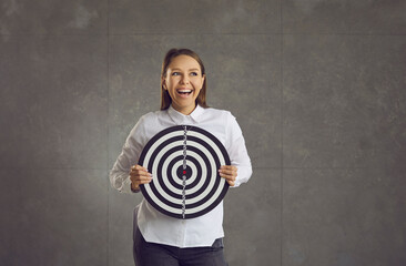 Young loudly laughing emotional caucasian woman holding dart board target standing on studio wall and looking aside. Business startup or project success, strategy planning, targeting marketing concept