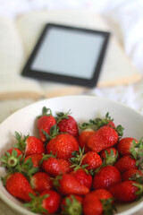 Bowl of strawberries, open book and e-reader on a bed. Selective focus.