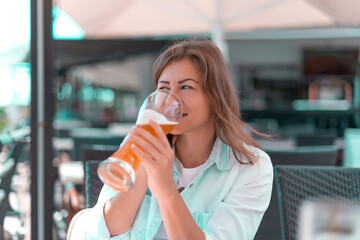 Young woman in jeans shirt is drinking beer on the restaurant terrace and talking by mobile phone