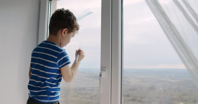Little boy with curly hair draws on the window at home with crayons. 