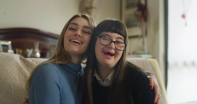 Authentic shot of happy young woman embracing her teen sister with down syndrome and smiling in camera at home. Concept of love, family, handicapped, healthcare, disability, love, friendship