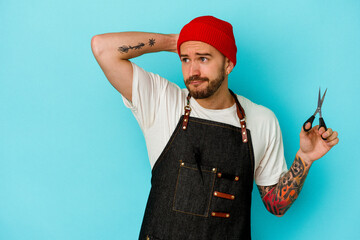 Young tattooed barber man isolated on blue background touching back of head, thinking and making a choice.